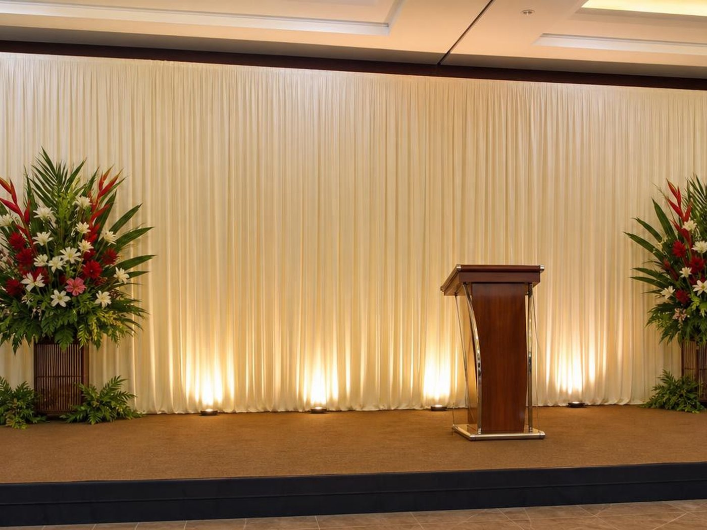 Backdrop panggung kain plisket ivory dengan uplight hangat dan podium di hall acara