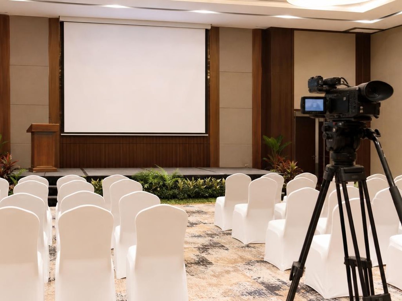 Setup seminar hybrid di ballroom hotel dengan layar kosong dan kamera video di baris belakang
